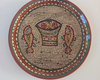 May include: A round ceramic plate with a mosaic design featuring two stylized fish flanking a central basket-like motif. The plate has a brown rim with a scalloped edge.