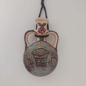 Könnte beinhalten: Eine kleine, cremefarbene Keramikflasche mit einer schwarzen Schnur, die oben hängt. Die Flasche hat zwei Henkel und ist mit einem Mosaikmuster und einer roten Blume verziert.