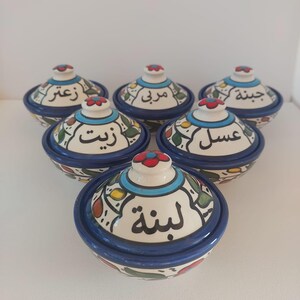 Puede incluir: Conjunto de seis pequeños cuencos de cerámica blanca con diseños azules y florales. Cada cuenco tiene una tapa y está etiquetado con escritura árabe. Las etiquetas dicen "Zait", "Hib", "Kune", "Zib", "Usl" y "Lish".
