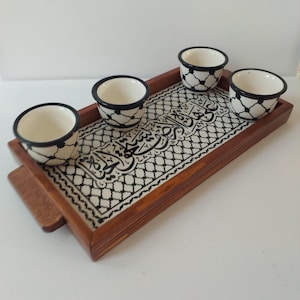 May include: A wooden tray with a black and white patterned design featuring Arabic calligraphy. The tray has four small white ceramic cups with black trim.