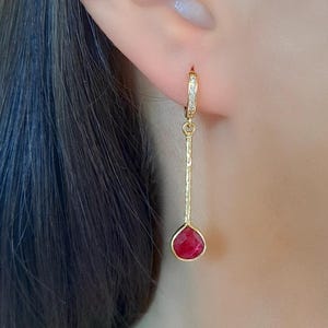 May include: A gold earring with a small hoop adorned with clear stones. A textured gold bar hangs from the hoop, ending in a teardrop-shaped, deep red gemstone. The earring is worn on a person's ear.
