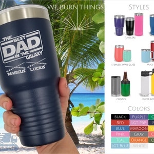 May include: A navy blue stainless steel tumbler with a white design that says "The Best Dad in the Galaxy" with two crossed lightsabers and the names "Marcus" and "Lucius" underneath. The tumbler is shown against a background of a beach and palm trees.
