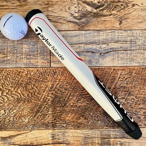 May include: White golf club grip with black and red accents. The grip has the text "TaylorMade" printed on it. A white golf ball is in the background.