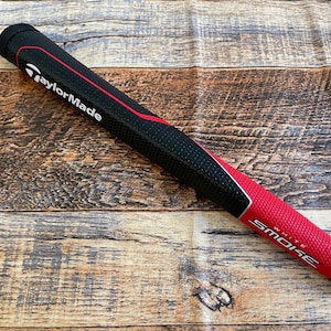 May include: Black and red golf putter grip with the text "TaylorMade" and "White Smoke" on it.