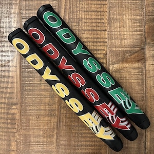 May include: Three black golf putter grips with the word "ODYSSEY" in yellow, red, and green lettering. The grips are angled on a wooden surface. The grips have a textured surface.
