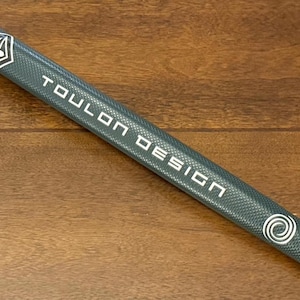 May include: A grey golf putter grip with a white "TOULON DESIGN" logo and a white swirl design at the end.