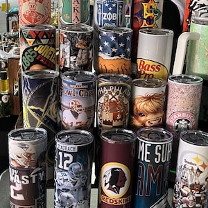 May include: A collection of insulated tumblers with diverse designs. Some tumblers display Greek letters, sports team logos, and other graphics. The colours include red, blue, green, and brown. The tumblers are cylindrical and appear to be made of stainless steel.