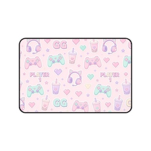 May include: A rectangular desk mat featuring a repeating pattern on a light pink background. The design includes pastel-coloured gaming controllers, cat-ear headphones, boba tea cups, hearts, stars, and the text "GG" and "PLAYER 1" in a soft font.