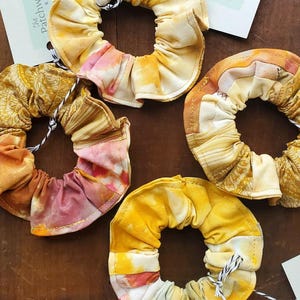 Puede incluir: Cuatro scrunchies de tela hechos a mano en tonos de amarillo, naranja y rosa. Los scrunchies están hechos de un patchwork de diferentes telas y tienen un efecto tie-dye. Los scrunchies se muestran sobre una superficie de madera con una etiqueta que dice "The Patchwork Palette".
