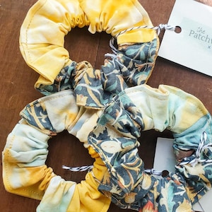 May include: Two handmade scrunchies with yellow and floral fabric. One scrunchie is primarily yellow with a tie-dye effect, while the other features a blue and green floral pattern. Each is attached to a white tag with the text "the Patchwork Palette".