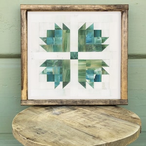 Op de afbeelding: Een ingelijst kunstwerk met een klaverbladontwerp in groen- en blauwtinten. Het kunstwerk wordt getoond op een rustieke houten kruk met een ronde bovenkant en drie poten. Het frame is gemaakt van donker hout.