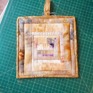 Puede incluir: Salvamanteles acolchado cuadrado con diseño de cabaña de troncos. El centro presenta un patchwork de telas naranja claro, morado y crema. El borde y la presilla para colgar son de tela dorada estampada. El salvamanteles está sobre una esterilla de corte verde.