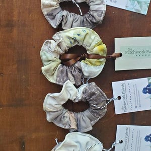 Puede incluir: Cinco scrunchies de tela hechos a mano en varios tonos de marrón, blanco y beige. Cada scrunchie tiene un diseño de patchwork único y está atado con una cinta marrón. Los scrunchies se muestran sobre una superficie de madera con una pequeña tarjeta adjunta a cada uno que dice "The Patchwork Palette".