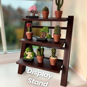 以下が含まれることがあります： 3 段の棚が付いた茶色の木製ディスプレイスタンド。各棚には、小さな鉢植えの植物が置かれています。植物には、サボテン、多肉植物、盆栽の木が含まれています。スタンドの下段には、「Display Stand」という文字が印刷されています。