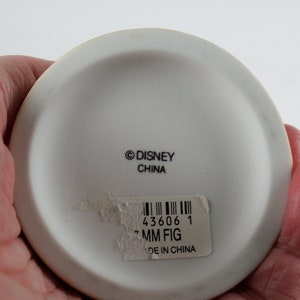 K&ouml;nnte beinhalten: Wei&szlig;e Keramik-Figuren-Basis mit dem Text "&copy;DISNEY CHINA" und einem Aufkleber mit einem Barcode, der Nummer "43606 1", dem Text "MM FIG" und "MADE IN CHINA".