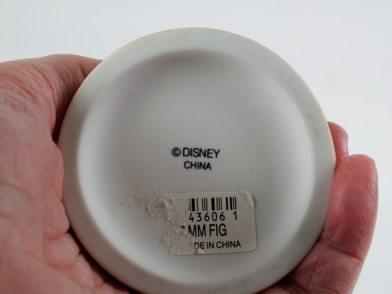 K&ouml;nnte beinhalten: Wei&szlig;e Keramik-Figuren-Basis mit dem Text "&copy;DISNEY CHINA" und einem Aufkleber mit einem Barcode, der Nummer "43606 1", dem Text "MM FIG" und "MADE IN CHINA".