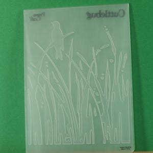 May include: A clear, rectangular embossing folder with a nature-themed design. The design features a bird perched on a branch, tall grass, and the word "Cuttlebug" at the top. The words "Paper Arts" are also visible.