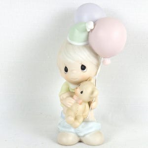 May include: A ceramic figurine of a child holding a teddy bear and two balloons. The child wears a green shirt, blue pants, and a green hat. The balloons are pink and lavender. The figurine has a sweet, innocent expression.