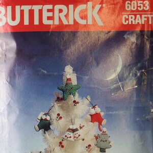 May include: A white Christmas tree decorated with handmade ornaments. The ornaments include a green star, snowmen, and red and green shapes. The background is a blue and white sky with a crescent moon. The top of the image reads "BUTTERICK 6053 CRAFT".