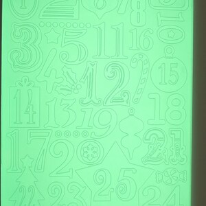 May include: A green Cuttlebug die-cut sheet featuring numbers and holiday-themed shapes. Includes numbers 1-25, Christmas tree, ornaments, and candy cane designs. The Provo Craft logo is visible in the upper right corner.