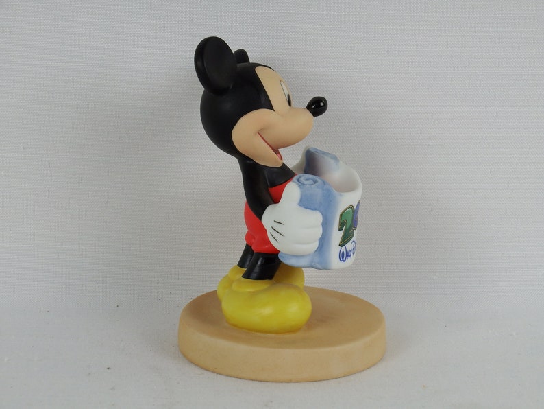 K&ouml;nnte beinhalten: Eine Keramikfigur von Mickey Mouse, die eine blaue und wei&szlig;e Schriftrolle mit dem Text "20th Anniversary Walt Disney World" h&auml;lt. Mickey tr&auml;gt seine charakteristischen roten Shorts und gelben Schuhe.