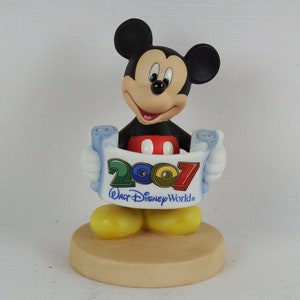 K&ouml;nnte beinhalten: Eine Keramikfigur von Mickey Mouse, die eine Schriftrolle mit dem Text "2007 Walt Disney World" h&auml;lt. Mickey tr&auml;gt seine charakteristischen roten Shorts und gelben Schuhe. Die Figur steht auf einem beigefarbenen Sockel.
