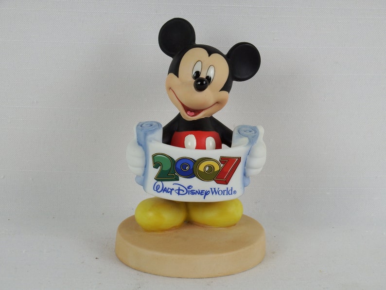 K&ouml;nnte beinhalten: Eine Keramikfigur von Mickey Mouse, die eine Schriftrolle mit dem Text "2007 Walt Disney World" h&auml;lt. Mickey tr&auml;gt seine charakteristischen roten Shorts und gelben Schuhe. Die Figur steht auf einem beigefarbenen Sockel.