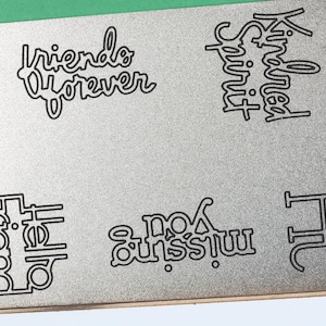 May include: A silver metal die cut with the phrases "friends forever", "kindred spirit", "hello friend", and "missing you" in a black, stylized font. The words are cut out of the metal. The words are for crafting and scrapbooking.