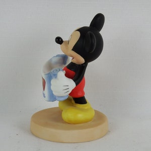 K&ouml;nnte beinhalten: Eine Keramikfigur von Mickey Mouse, die eine blaue und wei&szlig;e Karte mit einem roten Herzen h&auml;lt. Die Figur steht auf einem braunen Sockel.