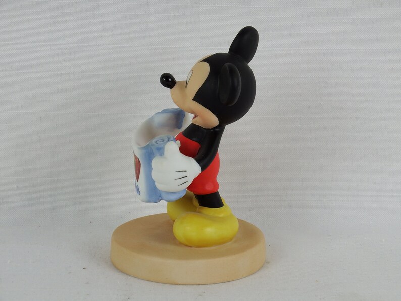 K&ouml;nnte beinhalten: Eine Keramikfigur von Mickey Mouse, die eine blaue und wei&szlig;e Karte mit einem roten Herzen h&auml;lt. Die Figur steht auf einem braunen Sockel.