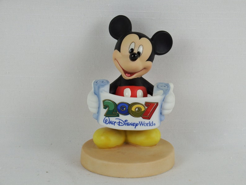 K&ouml;nnte beinhalten: Eine Keramikfigur von Mickey Mouse, die eine Schriftrolle mit dem Text "2007 Walt Disney World" h&auml;lt. Mickey tr&auml;gt seine charakteristischen roten Shorts und gelben Schuhe.