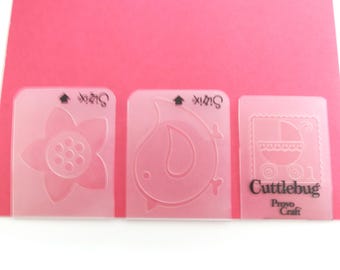 May include: Three pink Sizzix Cuttlebug embossing folders with flower, bird, and baby carriage designs. The baby carriage folder has the text "Cuttlebug Provo Craft". The folders are on a pink background.