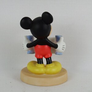 K&ouml;nnte beinhalten: Eine Keramikfigur von Mickey Mouse, die ein blaues und wei&szlig;es Schild mit erhobenen Daumen h&auml;lt. Die Figur steht auf einem braunen Sockel.