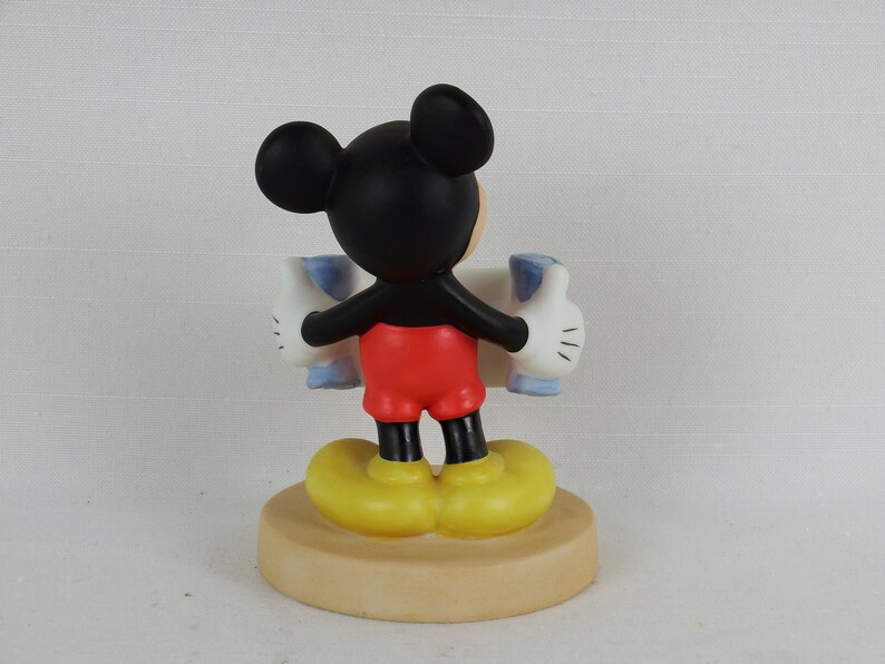 K&ouml;nnte beinhalten: Eine Keramikfigur von Mickey Mouse, die ein blaues und wei&szlig;es Schild mit erhobenen Daumen h&auml;lt. Die Figur steht auf einem braunen Sockel.