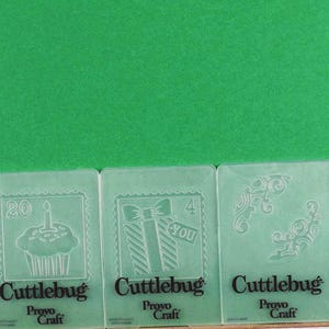 May include: Five clear plastic Cuttlebug embossing folders with various designs. The designs include a wreath, a cupcake with a candle, a gift with a bow, and decorative flourishes. The text "Cuttlebug" and "Provo Craft" are visible.