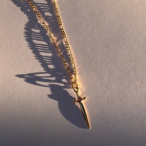 Könnte beinhalten: Goldfarbene Halskette mit einem Schwertanhänger. Der Anhänger hat einen roten Edelstein am Griff. Die Kette ist im feingliedrigen Stil gehalten. Die Halskette wird auf einer weißen Oberfläche präsentiert, mit einem Schatten nach links.