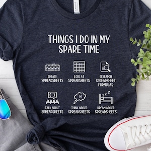 Może przedstawiać: Koszulka w kolorze ciemnoniebieskim z białym napisem "Things I do in my spare time". Tekst jest otoczony ikonami przedstawiającymi czynności związane z arkuszami kalkulacyjnymi. Koszulka leży na drewnianej powierzchni z dżinsami, okularami przeciwsłonecznymi i białymi butami sportowymi.