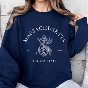 Puede incluir: Una sudadera azul marino con un gráfico blanco de un ancla, espadas cruzadas y un tridente. El texto "MASSACHUSETTS EST 1788 THE BAY STATE" está impreso en la sudadera.