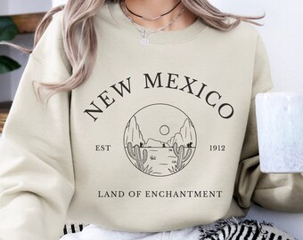 Könnte beinhalten: Sandfarbenes Sweatshirt mit einem kreisförmigen Grafikdesign mit dem Text "NEW MEXICO" und "LAND OF ENCHANTMENT". Die Grafik zeigt Kakteen, Berge und eine Sonne. Das Sweatshirt hat auch den Text "EST 1912."