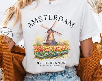 May include: White t-shirt with a watercolour illustration of a windmill in Amsterdam, Netherlands, surrounded by tulips. The word "AMSTERDAM" is arched above the image, with the country's name and coordinates below.