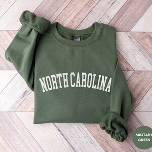 May include: A green sweatshirt with the text "NORTH CAROLINA" in white lettering. The sweatshirt is folded and laid on a woodgrain surface. The text "MILITARY GREEN" is visible in the lower right corner of the image.
