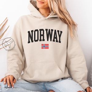 Op de afbeelding: Een zandkleurige hoodie met het woord "NORWAY" in vette zwarte letters over de borst, samen met de Noorse vlag. De hoodie heeft een voorzak en een capuchon met trekkoord. Het model draagt een lichtblauwe jeans.