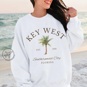 May include: White sweatshirt with the text "KEY WEST" in brown arched above a watercolor image of a palm tree. Below the tree are the words "EST. 1828" and "Southernmost City FLORIDA". The sweatshirt is a casual style.