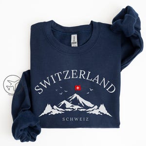 May include: Navy blue sweatshirt with the word "SWITZERLAND" in white arched above a mountain graphic. The Swiss flag is depicted on the mountain peak. The word "SCHWEIZ" is below the mountain graphic. The sweatshirt is a crew neck.