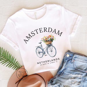 Może przedstawiać: Biała koszulka z napisem "AMSTERDAM" w łuku. Poniżej ilustracja roweru z koszem kwiatów. Tekst "NETHERLANDS" i współrzędne znajdują się poniżej. Szorty dżinsowe i kapelusz leżą obok koszulki.