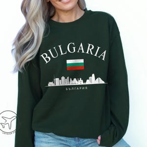 May include: Dark green sweatshirt with the word "BULGARIA" in white, above a Bulgarian flag and a white skyline graphic. The word "БЪЛГАРИЯ" is printed below the skyline. The sweatshirt is a crew neck style.