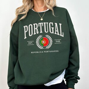 Puede incluir: Sudadera verde oscuro con la palabra "PORTUGAL" en blanco. Debajo, un emblema circular con la bandera portuguesa, el texto "EST. 1143" y "REPÚBLICA PORTUGUESA". La sudadera tiene mangas largas.