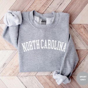 May include: A gray sweatshirt with the text "NORTH CAROLINA" in white, arched letters. The sweatshirt is folded and laid on a woodgrain surface. The text "SPORT GREY" is in the lower right corner of the image.