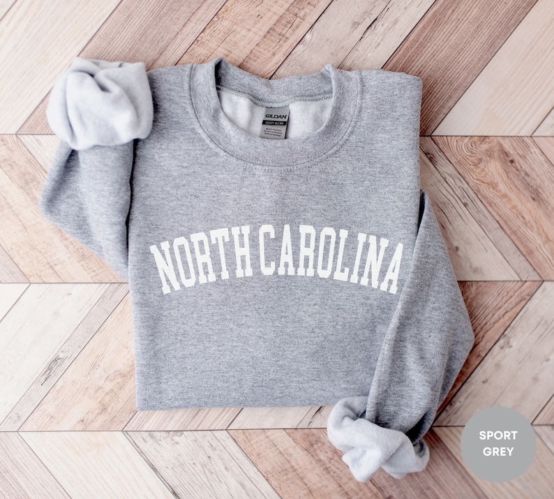 May include: A gray sweatshirt with the text "NORTH CAROLINA" in white, arched letters. The sweatshirt is folded and laid on a woodgrain surface. The text "SPORT GREY" is in the lower right corner of the image.