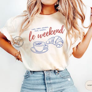 May include: Ivory-colored t-shirt with the phrase "le weekend" in red script, accompanied by a blue line drawing of a coffee cup and croissant. The shirt also features the days of the week in French.